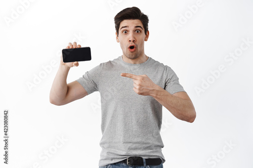 Impressed guy pointing at cool app, empty smartphone screen, showing moible phone display, standing against white background photo