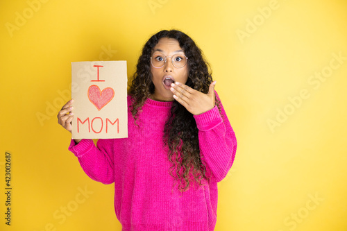 Beautiful woman celebrating mothers day holding poster love mom message covering mouth with hand, shocked and afraid for mistake. surprised expression