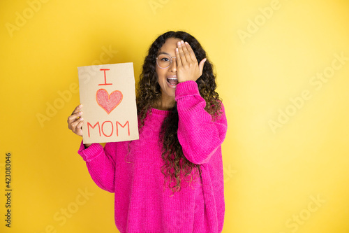 Beautiful woman celebrating mothers day holding poster love mom message covering one eye with hand, confident smile on face and surprise emotion.