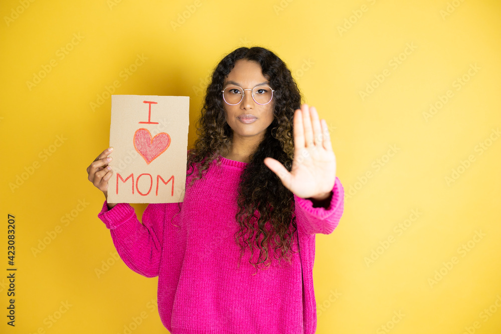 Beautiful woman celebrating mothers day holding poster love mom message serious and doing stop sing with palm of the hand. Beautiful woman celebrating mothers day holding poster love mom message serious and doing stop sing with palm of the hand.