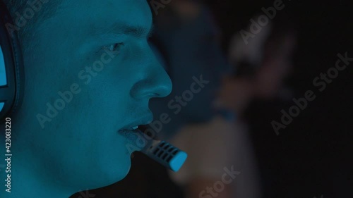 Portrait of a man speaking into a microphone playing a video game. It is illuminated by a monitor with overflowing colors. He plays with his team that sits next to. High quality 4k footage