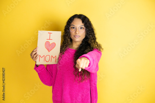 Beautiful woman celebrating mothers day holding poster love mom message pointing with finger to the camera and to you, confident gesture looking serious
