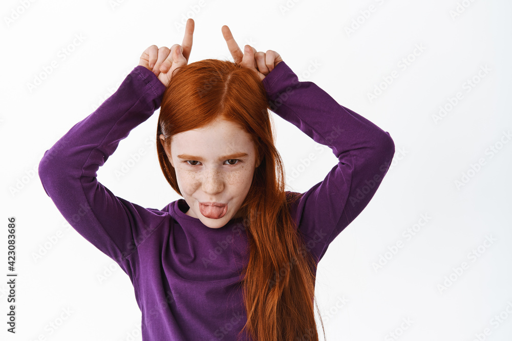 Beautiful redhead little kid girl shows bull horns and tongue, smiling sly at camera, playing and having fun, standing over white background Beautiful redhead little kid girl shows bull horns and tongue, smiling sly at camera, playing and having fun, standing over white background