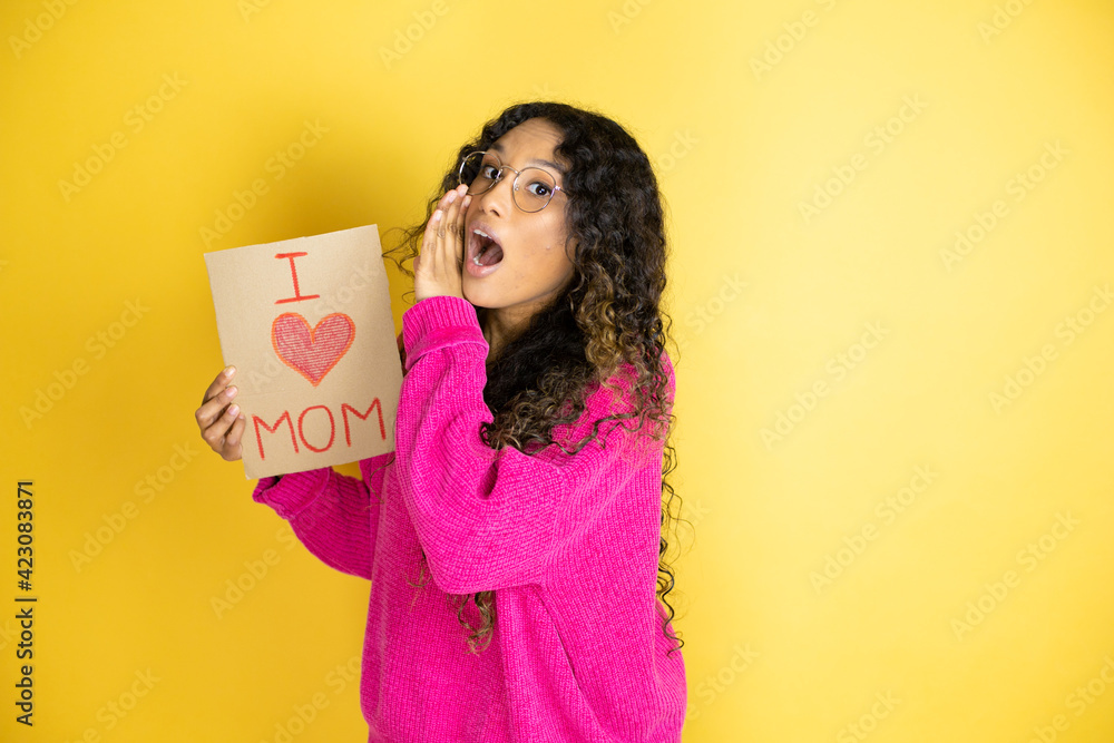 Beautiful woman celebrating mothers day holding poster love mom message with hand on mouth telling secret rumor, whispering malicious talk conversation Beautiful woman celebrating mothers day holding poster love mom message with hand on mouth telling secret rumor, whispering malicious talk conversation