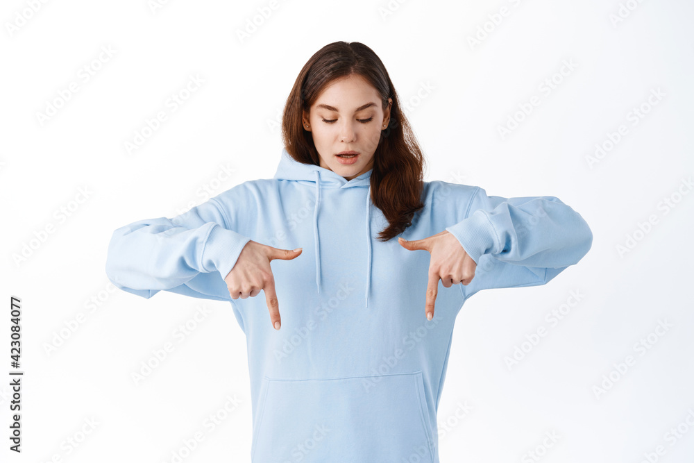 Young brunette teen girl pointing and looking down with amazed gasp, showing promo logo on bottom copy space, staring excited, standing over white background Young brunette teen girl pointing and looking down with amazed gasp, showing promo logo on bottom copy space, staring excited, standing over white background