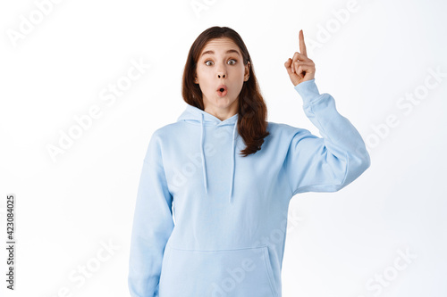 Surprised and excited brunette woman model, gasping amazed, pointing finger up at promo logo, showing advertisement, standing against white background