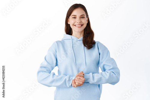 Cute upbeat girl ready to help you, holding hands together and smiling friendly. Young woman standing confident and happy, gladly answer your questions, standing over white background