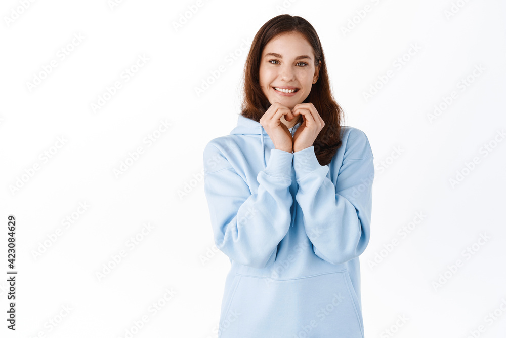 Cute young woman in hoodie, showing hand heart gesture and smiling at you, love and like sign, standing against white background Cute young woman in hoodie, showing hand heart gesture and smiling at you, love and like sign, standing against white background