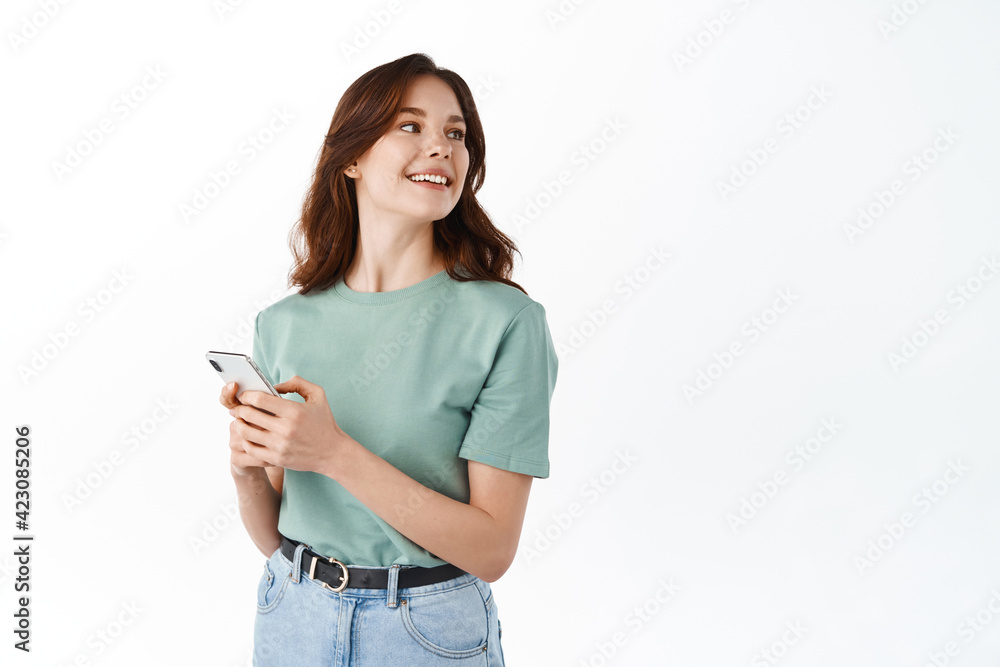 Young modern girl standing with smartphone and look aside at copy space, smiling while chatting on social media app, standing against white background Young modern girl standing with smartphone and look aside at copy space, smiling while chatting on social media app, standing against white background