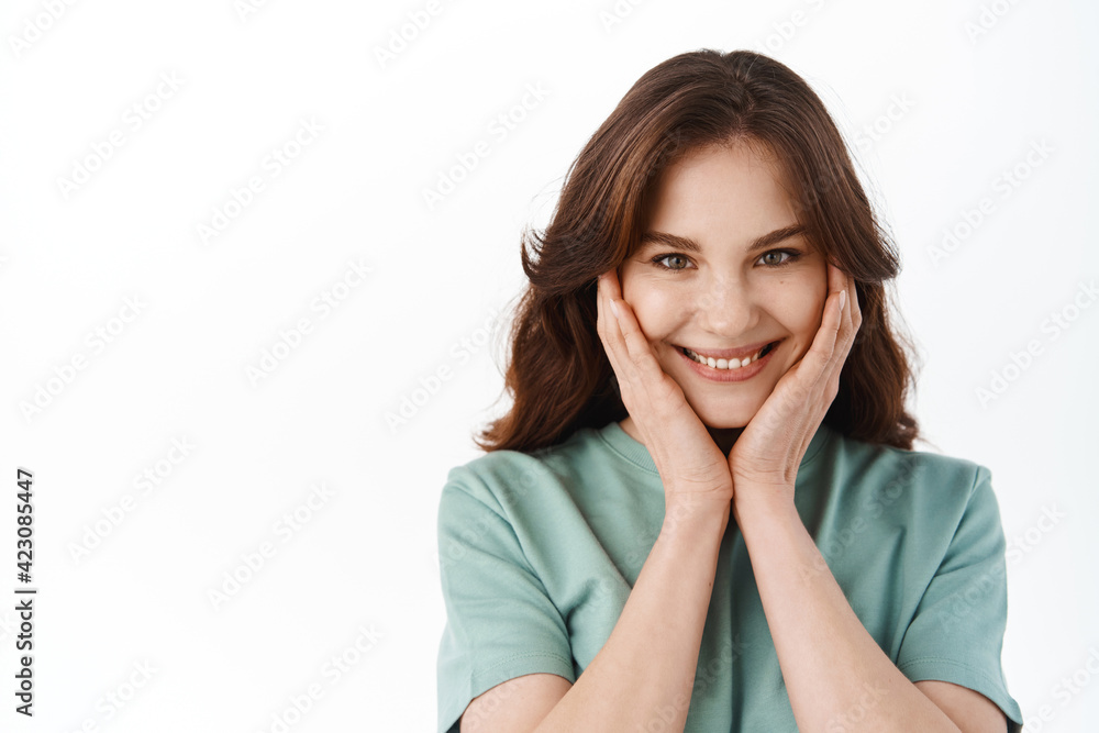 Beautiful girl smiling and blushing, touching her cheeks and looking flattered, receive compliments or praises, standing against white background Beautiful girl smiling and blushing, touching her cheeks and looking flattered, receive compliments or praises, standing against white background