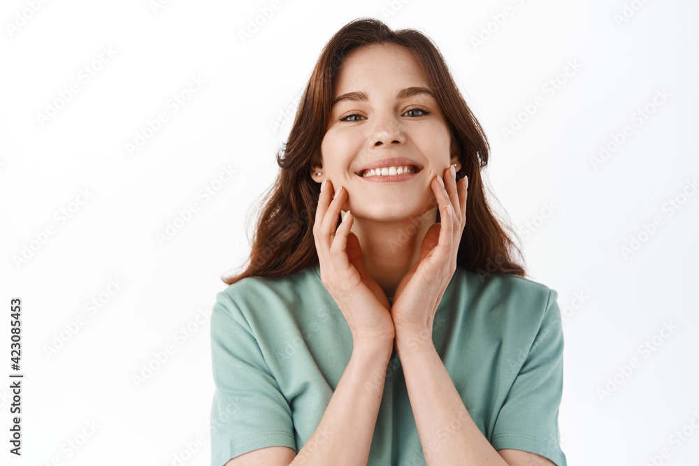 Skin care. Beautiful teen girl touching her perfect clean face and smiling pleased, using cosmetics for better face hydration and nourish effect, standing over white background Skin care. Beautiful teen girl touching her perfect clean face and smiling pleased, using cosmetics for better face hydration and nourish effect, standing over white background