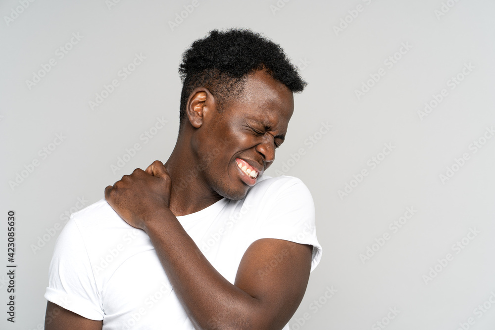Unhappy Black man suffering from neck pain, pinched nerve in her back. African male has spinal problem, shoulder stiffness, muscle inflammation, rheumatoid pain isolated on studio grey background. - obrazy, fototapety, plakaty 