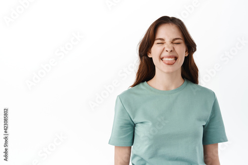 Happy beautiful girl having fun, being herself and showing true emotions, stick tongue and squinting joyful at camera, standing against white background © Cookie Studio