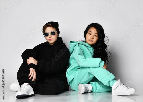 Trendy fashion interracial sportive boy and girl sitting on studio floor. Full-length portrait of caucasian male and asian female children wearing warm sportswear. Teenage kid vogue and style concept