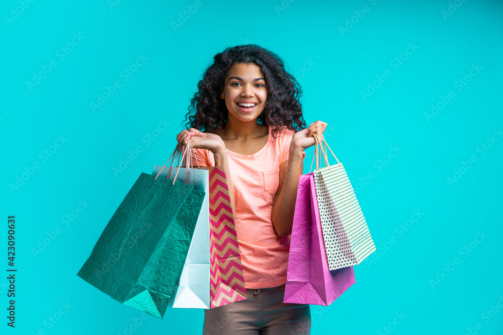 Cute excited african american girl in casual outfit posing with piles of paper shopping bags isolated over blue background