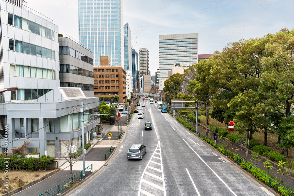 Obraz premium Urban road lined with office and residential buildings on one side and with a park on the other side in central Tokyo, Japan, on a cloudy spring day