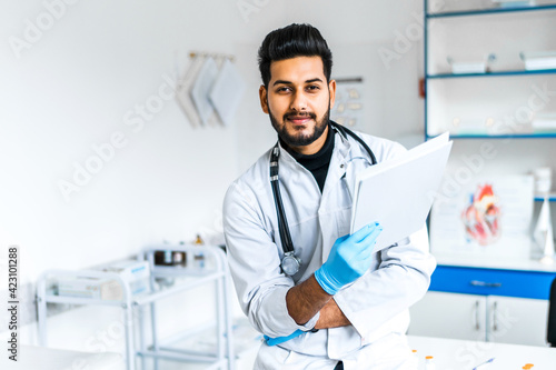 A handsome Indian doctor man in a medical suit in the middle of a modern hospital, he is looking at the camera