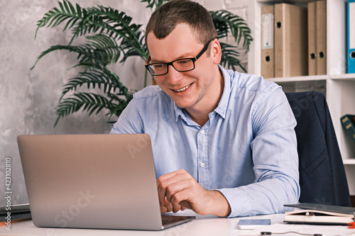 Cheerful smiling 30 year old man in glasses and a shirt looks into the laptop screen while sitting in a light office on the background of a rack with folders. Office work, entrepreneurship concept