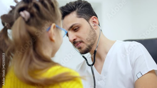 Portrait of handsome positive pediatrician listening auscultation of girl using stethoscope and talking. Cheerful Middle Eastern man consulting little Caucasian patient in hospital. Pediatric clinic.