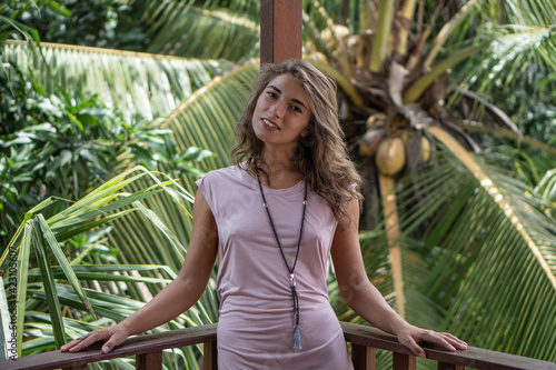 Portrait of young beautiful woman in pink dress on balcony of tropical bungalow with palm trees view photo