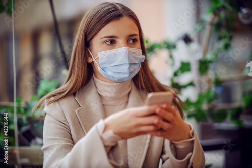 Portrait of girl with face mask on sitting in cafe outdoors and using phone for e-banking.