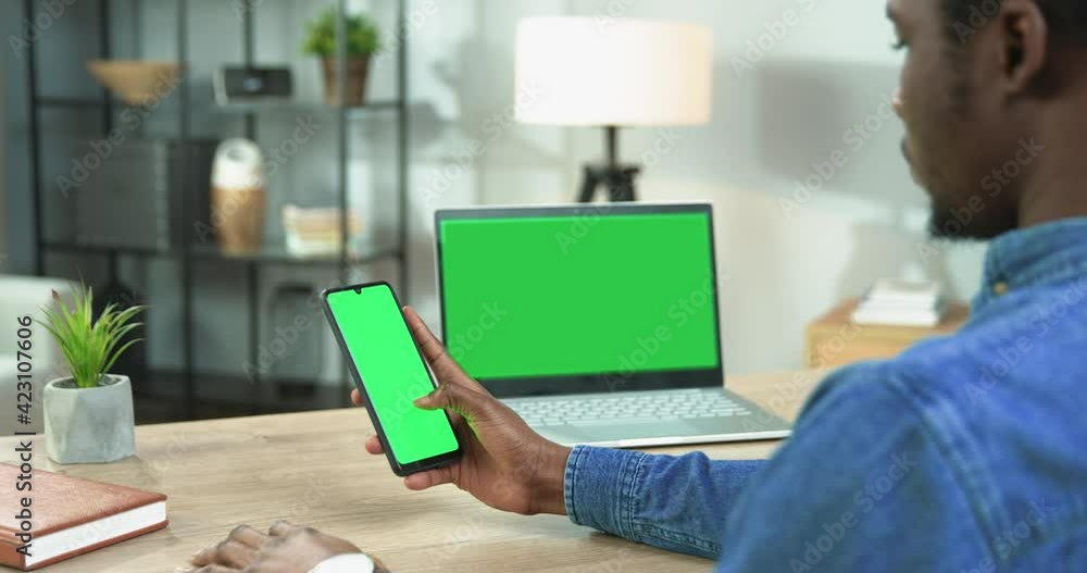 Close up of African American guy scrolling on smartphone with green screen browsing social network app while looking at laptop with chroma key sitting at desk in living room, leisure concept Close up of African American guy scrolling on smartphone with green screen browsing social network app while looking at laptop with chroma key sitting at desk in living room, leisure concept
