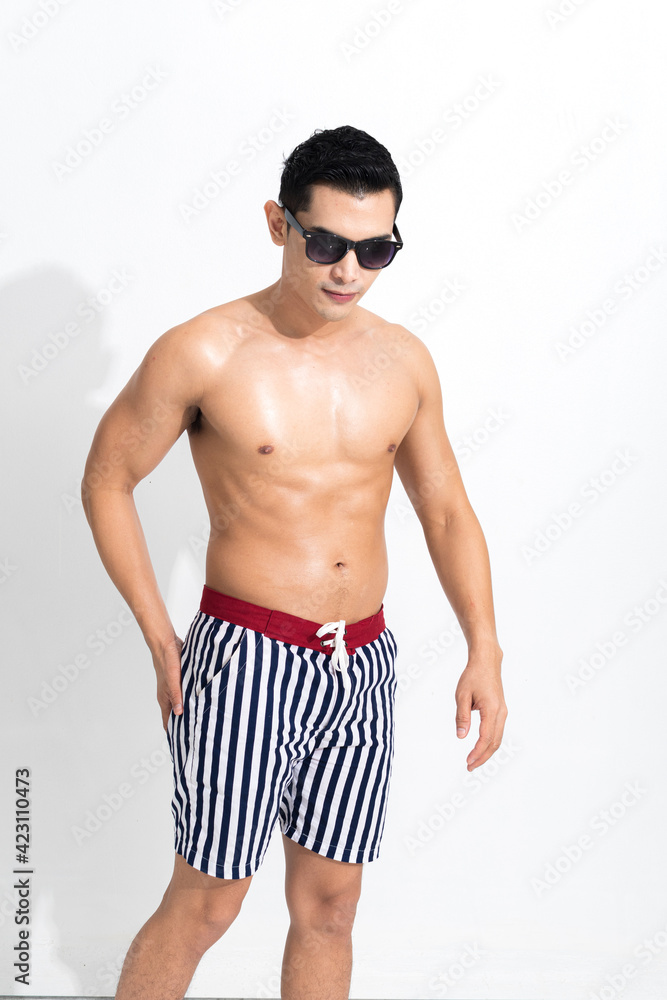 Young muscular guy in striped beach shorts sunbathing wear sunglasses at studio shot isolated on white background. Fashion summer concept.