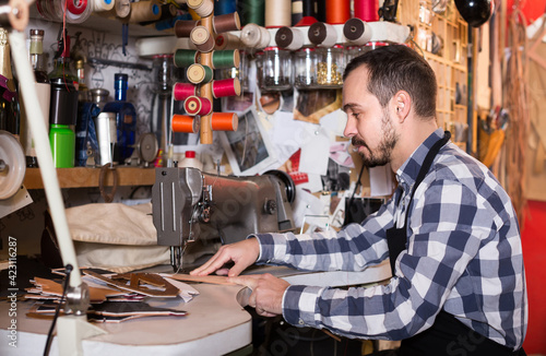 positive russian male worker sewing stitches on belt in leather workshop