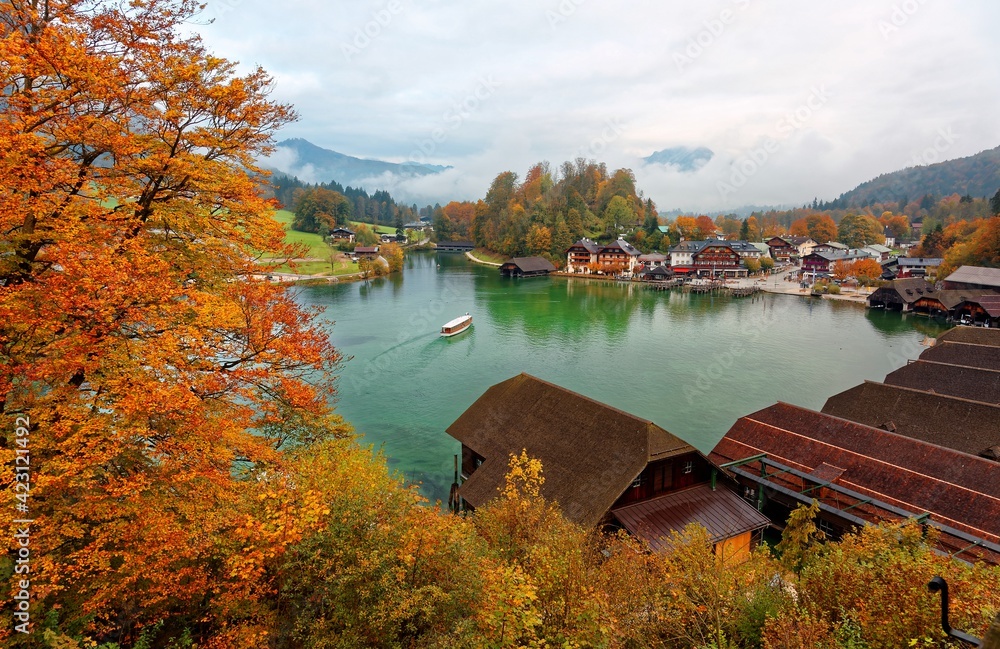 A sightseeing boat cruising on Konigssee ( King's Lake ) surrounded by colorful autumn trees and boathouses on a foggy misty morning~ Beautiful scenery of Bavarian countryside in Berchtesgaden Germany