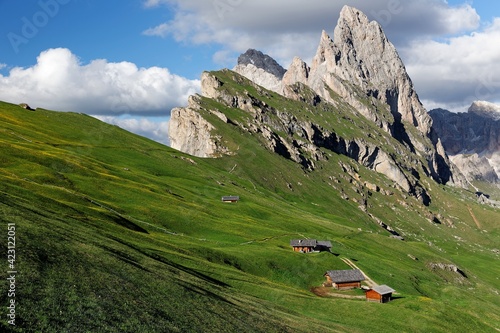 Beautiful scenery of Dolomites on a sunny summer day with view of majestic Odle (Geisler) mountain peaks in background & wooden huts on grassy hills at Seceda, Val Gardena, South Tyrol, Italy, Europe