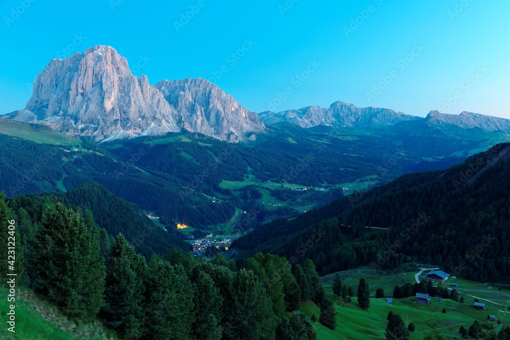 Scenery of majestic Sassolungo-Sassopiatto mountains at blue dusk & lights from village houses in the green grassy valley in Col Raiser, Val Gardena, Dolomiti National Park, South Tyrol, Italy, Europe