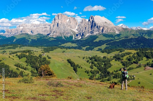 A tourist & his dog enjoying the majestic scenery of rugged Sassolungo-Sassopiatto & green grassy valley under sunny sky in Ortisei, Seiser Alm Highland (Alpe di Siusi), Dolomites, South Tyrol, Italy