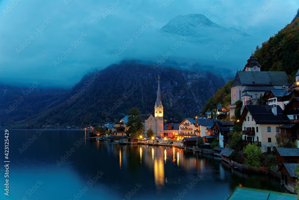 Morning view of Hallstatt, a peaceful lakeside village in Salzkammergut region of Austria, with majestic mountains & lights of village houses & the church reflected on lake water in a deep blue mood