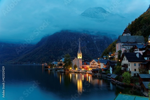 Morning view of Hallstatt, a peaceful lakeside village in Salzkammergut region of Austria, with majestic mountains & lights of village houses & the church reflected on lake water in a deep blue mood © AaronPlayStation