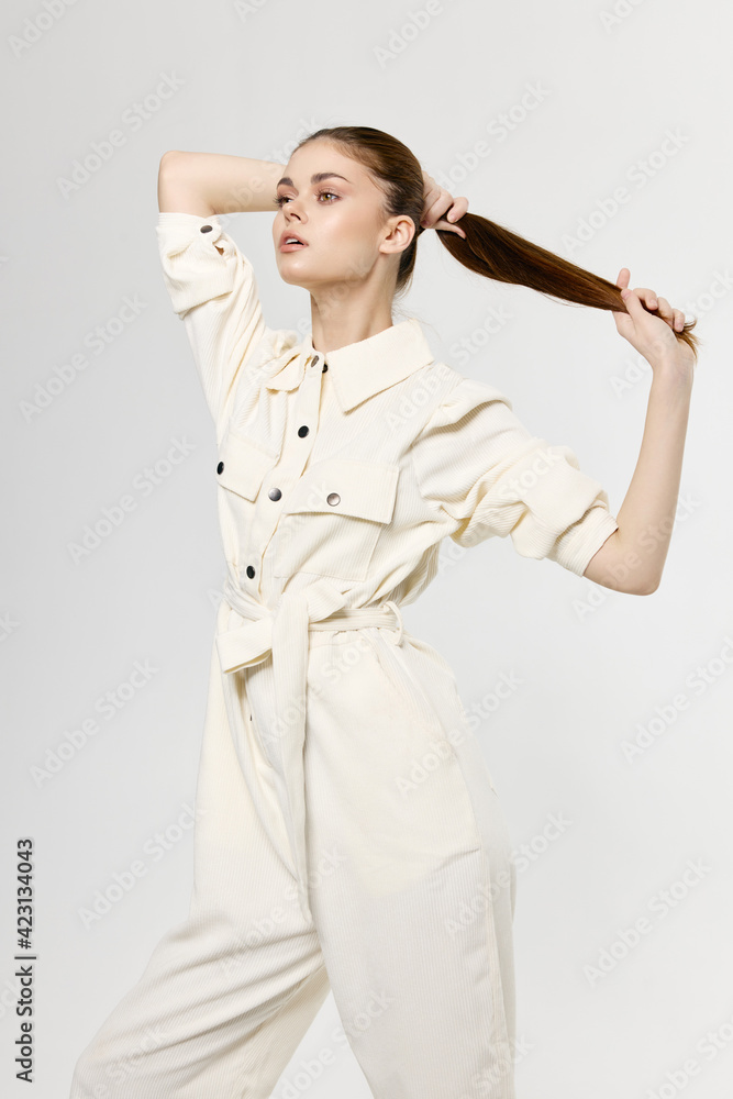 Woman in white jumpsuit touches hair on her head with her hands on a light background