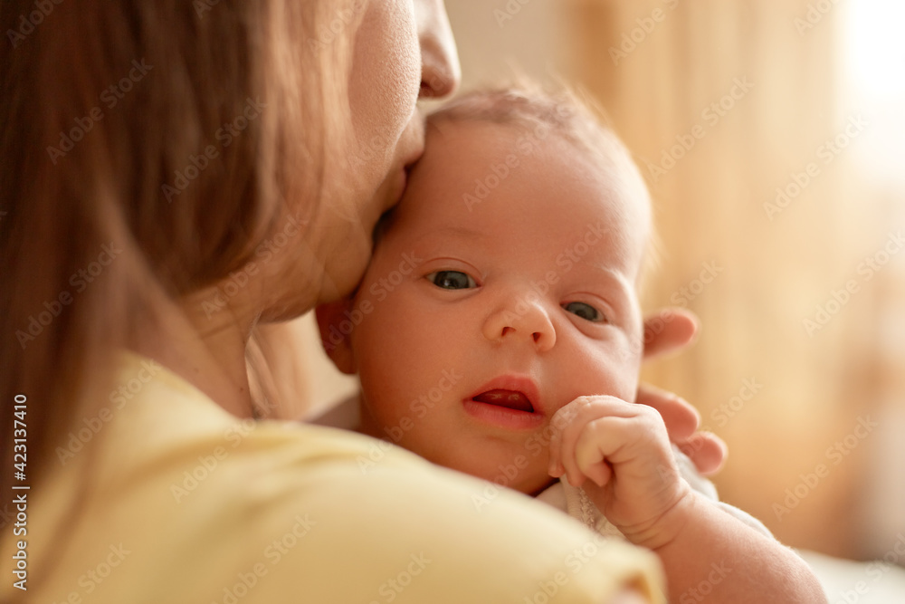 Mother posing backwards and holding newborn baby, mommy kissing tiny infant, kid looking at camera, keeping tiny fist near face, family posing indoor. Mother posing backwards and holding newborn baby, mommy kissing tiny infant, kid looking at camera, keeping tiny fist near face, family posing indoor.