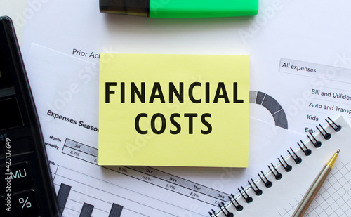 Text FINANCIAL COSTS on the page of a notepad lying on financial charts on the office desk.