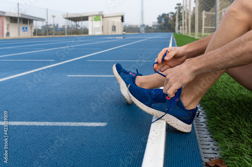 Unrecognized male athlete sitting in the grass in the running track tying his shoelaces.