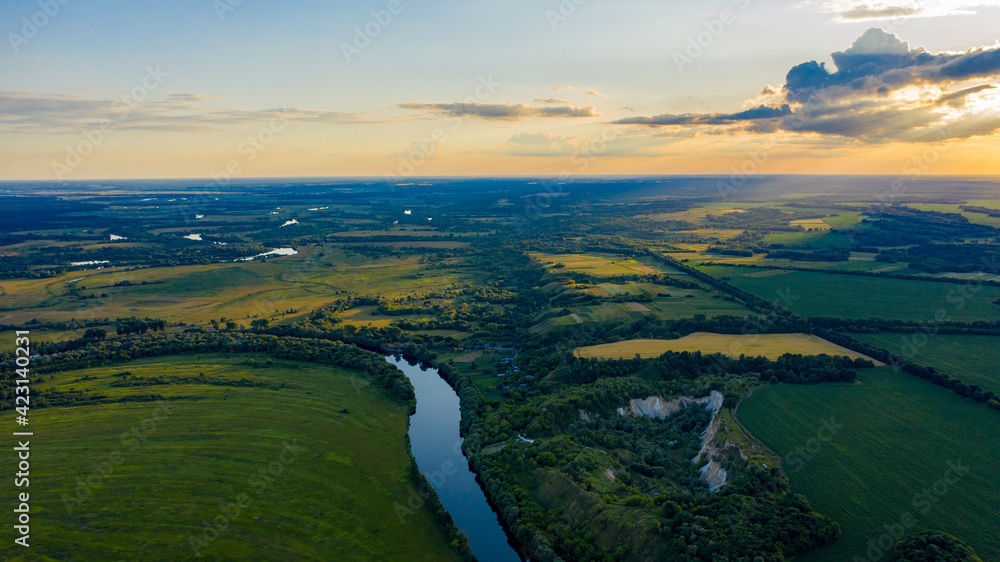 Fototapeta premium Aerial view of a beautiful summer hilly landscape with a river while sunset. Wide green valley with a river running in the middle. Green meadows. Top view over beauty nature.