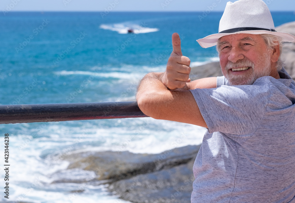 Portrait of carefree senior man in sea vacation, looking at camera and smiling with thumb up. Handsome retired enjoying relax