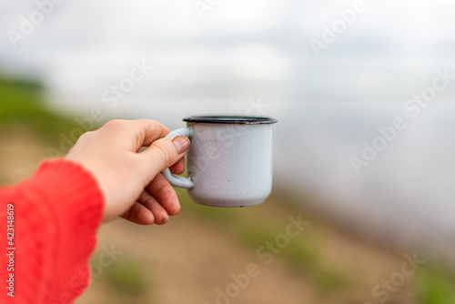 a female hand in a red sweater holds an iron mug against the background of the river and the sky. Outdoor recreation concept.