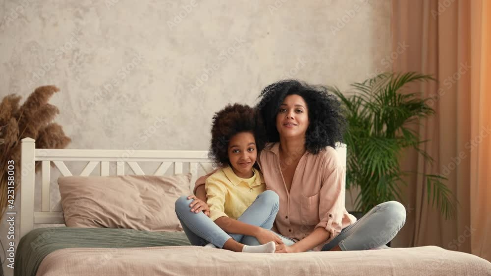 African American woman and little girl are hugging each other and have fun talking. Mother and daughter posing, sitting on the bed in bedroom with bright interior. Slow motion ready, 4K at 59.97 fps. African American woman and little girl are hugging each other and have fun talking. Mother and daughter posing, sitting on the bed in bedroom with bright interior. Slow motion ready, 4K at 59.97 fps.