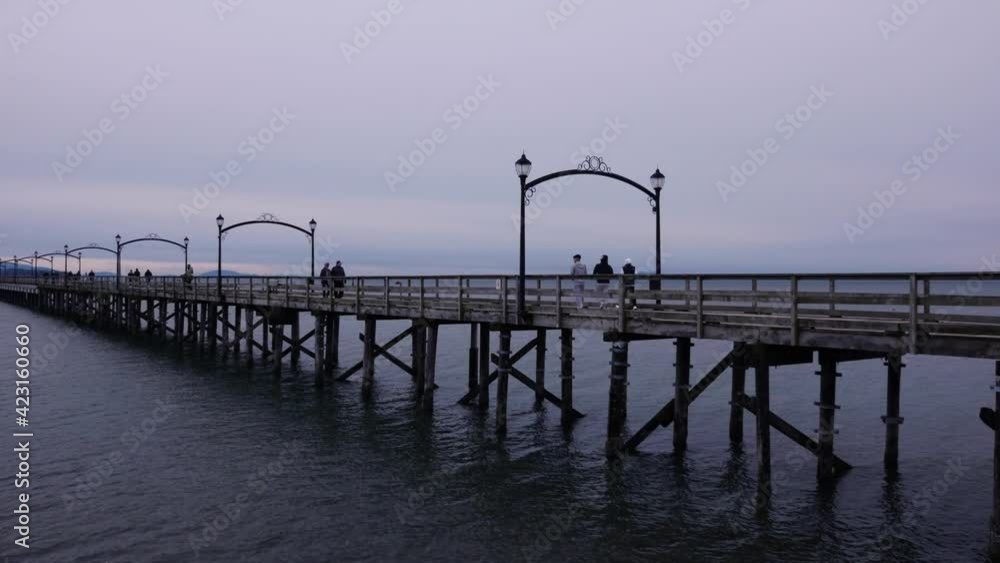 Static view of people walking on a long pier leading towards a break water in ocean during the evening clouds at dusk Static view of people walking on a long pier leading towards a break water in ocean during the evening clouds at dusk
