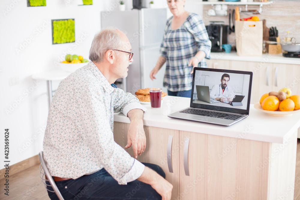 Senior retired man in video conference with doctor using laptop in kitchen. Online health consultation for elderly people drugs ilness advice on symptoms, physician telemedicine webcam. Medical care Senior retired man in video conference with doctor using laptop in kitchen. Online health consultation for elderly people drugs ilness advice on symptoms, physician telemedicine webcam. Medical care