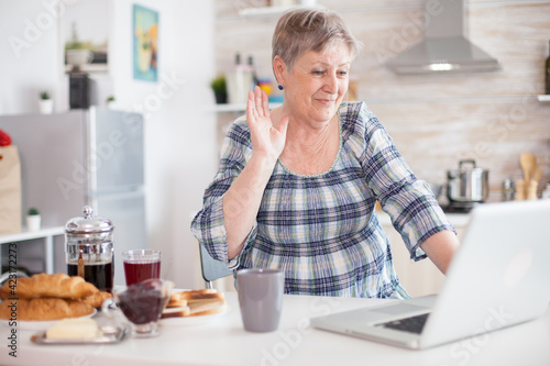 Senior woman drinking coffee and waving during video conference with family in kitchen while having breakfast. Elderly person using internet online chat technology video webcam making a video call