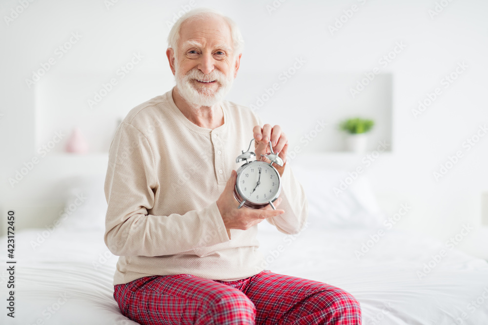 Photo of elderly man pensioner sit on bad home happy positive smile hold clock alarm timer wear pajamas Photo of elderly man pensioner sit on bad home happy positive smile hold clock alarm timer wear pajamas