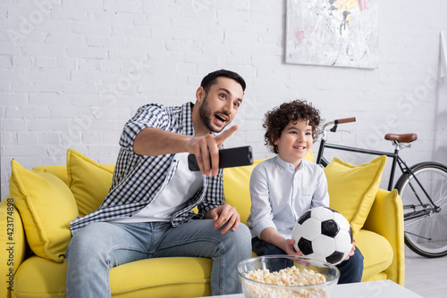 Arabian man with remote controller pointing with finger near popcorn and son with football at home