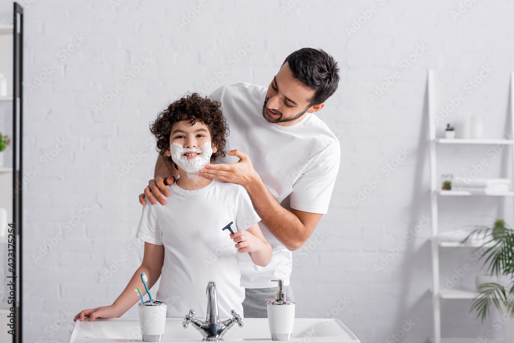 Smiling arabian father applying shaving foam on face of preteen son with razor in bathroom Smiling arabian father applying shaving foam on face of preteen son with razor in bathroom