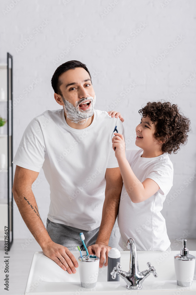 Arabian boy holding razor near smiling father in shaving foam in ...