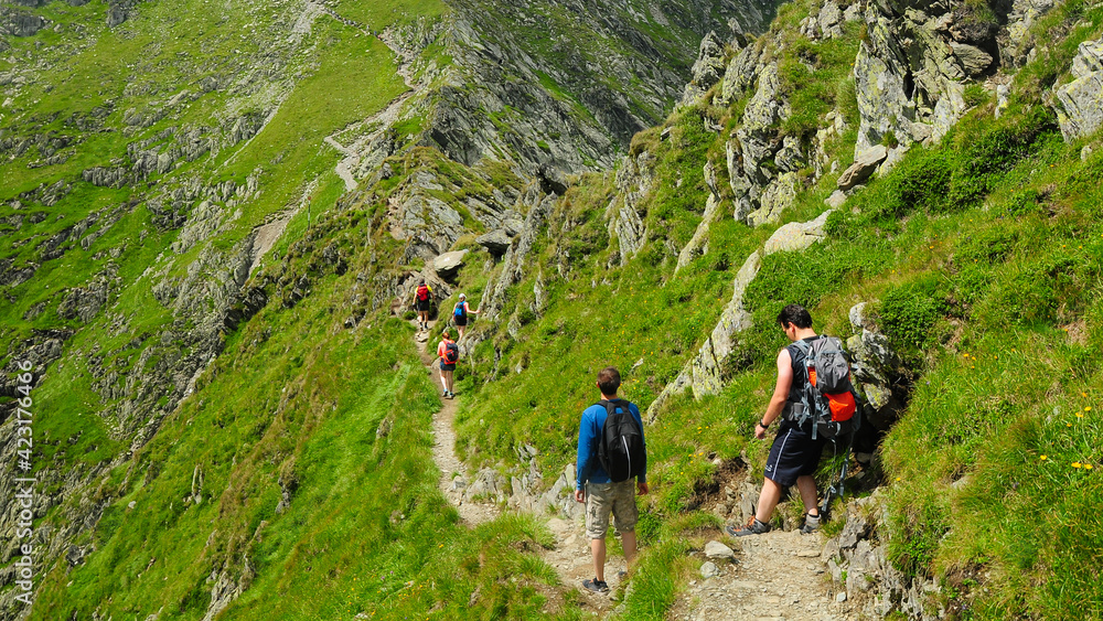 Hikers climbing on trail in Fagaras Mountains. The footpath winds along a steep rocky ridge, narrowed by an abyss. Carpathia, Romania.